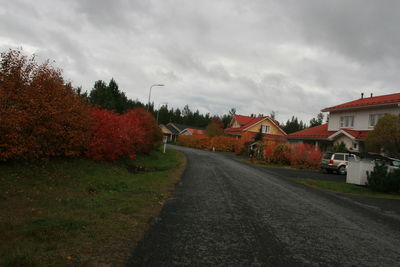 Empty road along houses and trees against cloudy sky