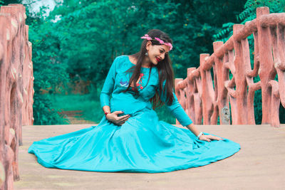 Pregnant woman wearing turquoise evening gown while sitting on footbridge against trees