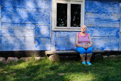 Senior woman sits on a bench in front of a wooden blue  house 