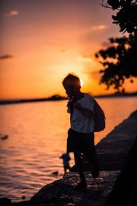 Silhouette boy standing on shore against orange sky