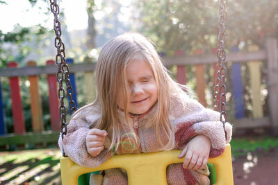 A young girl sits on a swing in a retro-styled playground, her expression is thoughtful. 