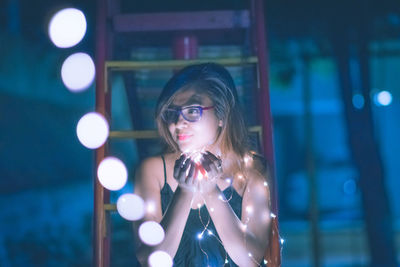 Beautiful woman holding illuminated string lights against steps at night