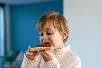 A boy bites into a sandwich with hazelnuts and cocoa, sitting on a table in the living room.