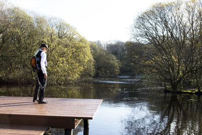 Full length of senior man with guitar standing on pier over lake against sky