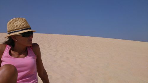 Young woman wearing hat while resting on sand against clear sky at beach