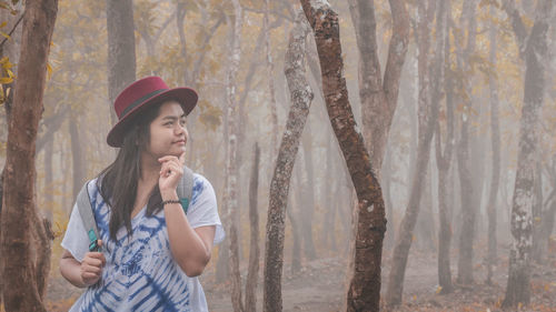 Woman with hand on chin standing in forest during foggy weather