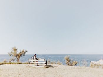 Rear view of man sitting on bench by sea against clear sky
