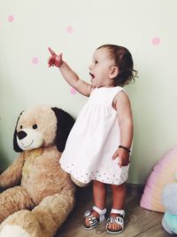 Cute baby girl pointing while standing by stuffed teddy bear against wall