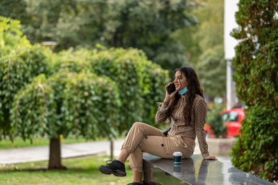 Attractive teen girl with smartphone and coffee sitting in a park and having a phone conversation. 