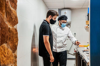 Male cook in medical mask talking to waiter and pointing at prepared delicious dish ready for serving in cafe