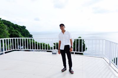 Full length portrait of young man standing on railing against sky