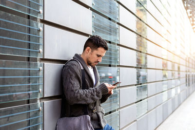 Side view of a bearded man using phone leaning on office building wall