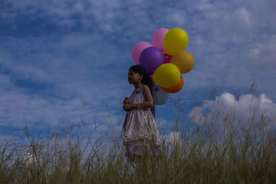 Girl holding balloons while standing by plants against sky