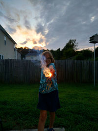 Happy girl holding illuminated sparkler in yard against sky