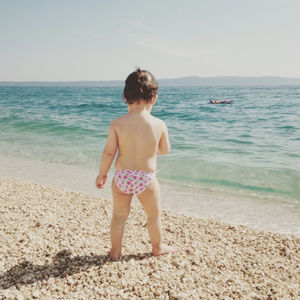 Full length rear view of baby standing at beach against sky during sunny day