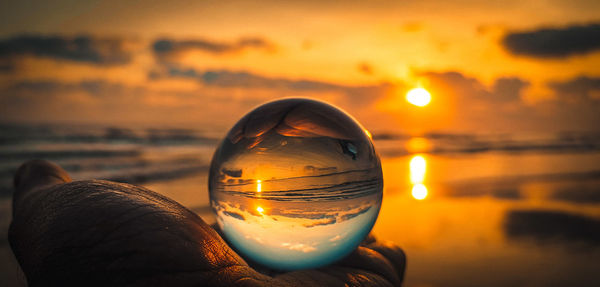 Close-up of person hand holding crystal ball against sea and sky during sunset