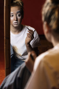 Young professional female mime in casual clothes painting the eyebrows and looking in mirror in dressing room