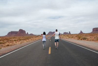 Full length rear view of couple holding hands while walking on country road at desert against cloudy sky