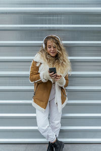 Young woman listening music through smart phone while standing against metal wall