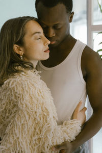 Young woman wearing fur jacket with eyes closed embracing boyfriend at home