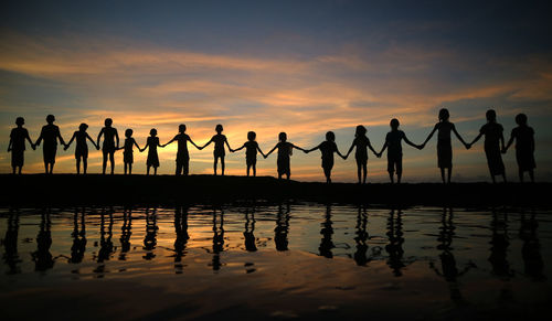 Silhouette children holding hands while standing against sky during sunset