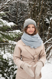 A cute girl is standing under the snow in the forest in mittens and a coat