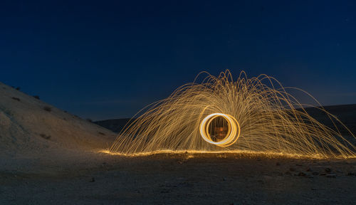 Person spinning wire wool while standing on sand against sky at night