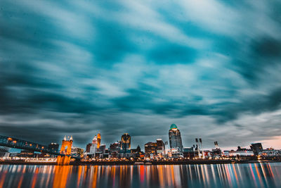 Illuminated city at waterfront against cloudy sky