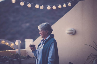 Senior woman holding wineglass while standing outdoors during sunset