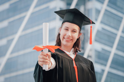 Portrait of young woman wearing graduation gown while standing against office building