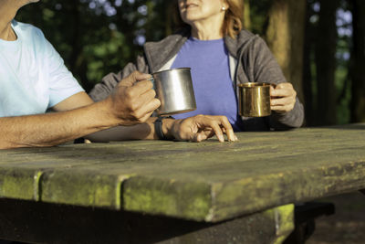 Unrecognizable senior couple holding and drinking from metal cups on a camping table while talking