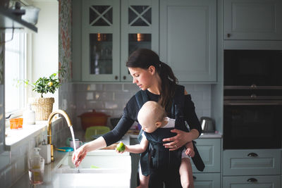 Mother washing hand in kitchen sink while carrying baby girl at home