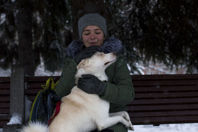 Young woman stroking dog while sitting on bench at park during winter