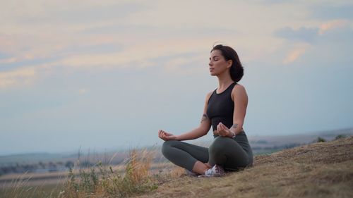 Side view of woman sitting on rock against sky during sunset