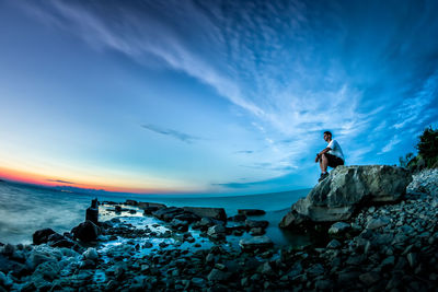 Side view of man sitting on rocks by sea against dramatic sky during sunset