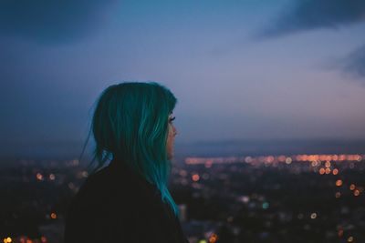 Side view of thoughtful woman standing against illuminated city at dusk