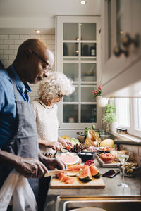 Multi-ethnic senior friends cutting fruits at counter while preparing dinner in kitchen