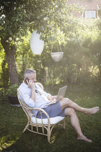 Full length of man talking on smart phone while using laptop in backyard