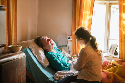 Female nurse feeding paralyzed elderly lady with puree through tube and syringe while taking care of patient at home