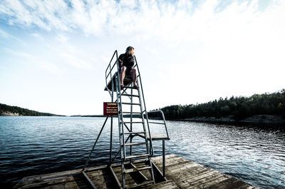 Woman sitting on diving platform by lake against sky