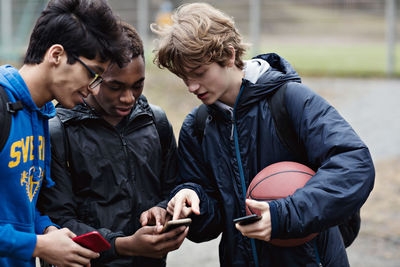 Friends looking in teenage boy's phone while standing with ball on playing field