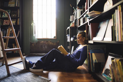 Side view of young woman reading book while reclining on shelf in bookstore