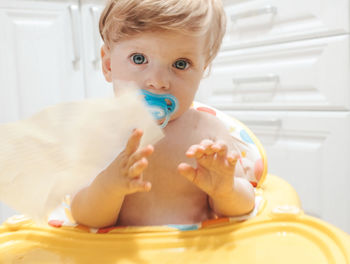 Close-up portrait of shirtless baby girl with pacifier sitting in high chair