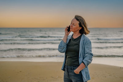 A young woman on the beach near the ocean in the spring at sunset talking on the phone with a smile