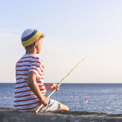 Boy sitting on retaining wall while fishing in sea