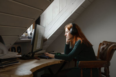 Middle age woman listening to music in big headphones near computer at home. creating play list