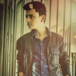 Fashionable teenage boy looking away while standing against glass wall