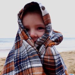 Portrait of child wrapped in blanket at beach against sky