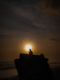 Silhouette person on rock by sea against sky during sunset