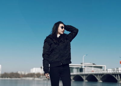 Woman wearing jacket standing by river against blue sky